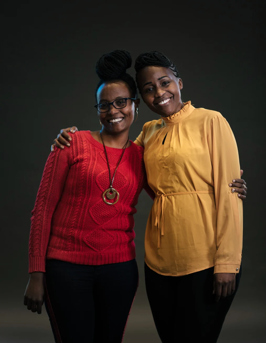 In this picture, two women with deep skin tones are shown smiling toward the camera in front of a black
backsplash. One woman has a red sweater, black pants, and glasses, while the other has a yellow 
drawcord waist shirt dress and black pants.