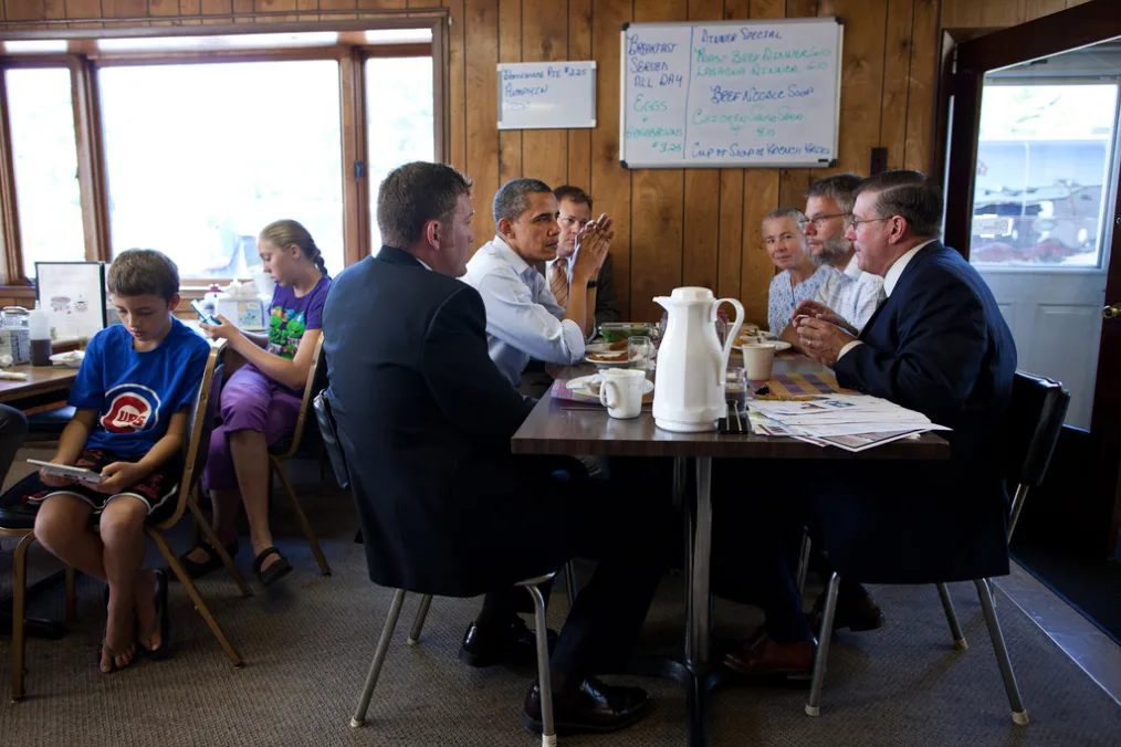 President Obama wearing a light blue button-up shirt sits on the left-hand side of a dark brown wooden table with five other individuals with light skin tones. There are two kids with light skin tones sitting at the table behind them. There is a wooden board wall with a large and small white board and a window. 
