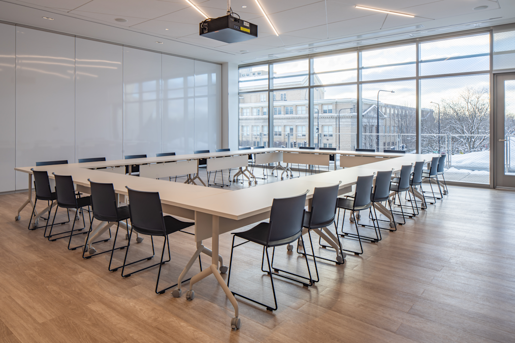 White tables and dark blue chairs are arranged in a large square inside a room with light wood floors. Large windows on the right look out onto a snowy street. A black projector hangs from the ceiling.