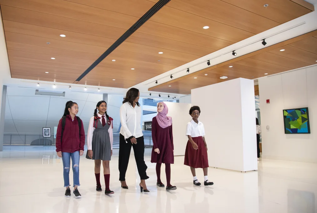 Michelle Obama walks with four young girls who each have different skin tones. Michelle Obama has a white, silk, long sleeve collared shirt, heels, and black pants on. The girls have red, white, grey, black, and blue businees casual themed attire on. They are walking through a wide white hallway. Art can be seen in the background.