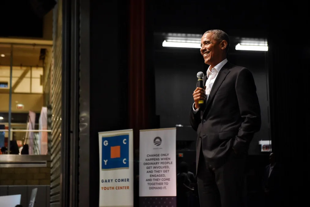 Barack Obama a black man standing on stage smiling while holding a mic and wearing a black and white suit