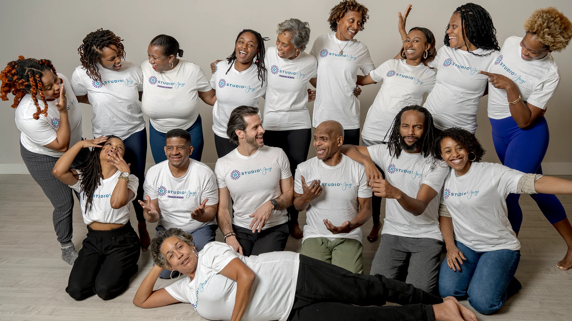 A group shot of Julia Perkins, a woman with a light medium skin tone, lays at the front of a group of people with a range of light to dark skin tones smiling and posing for a photo. They all wear white shirts that read, “Studio Yogi.”