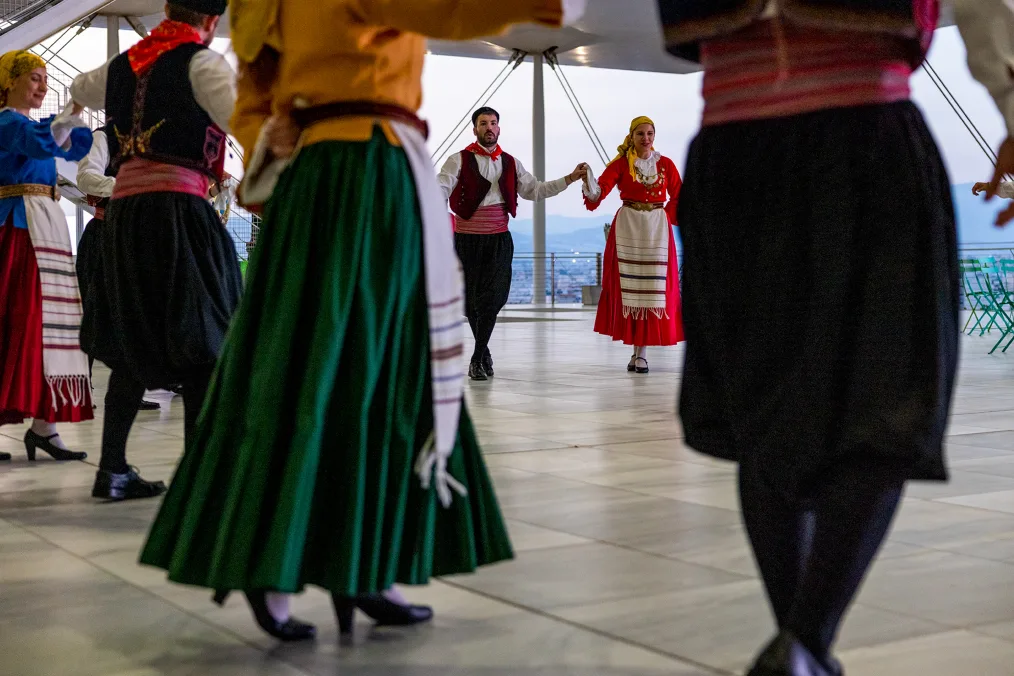 A group of people hold hands and form a circle to participate in a traditional Greek dance. Six men and women of varying skin tones are visible. They are wearing red, black, and green skirts in a range of vibrant colors.