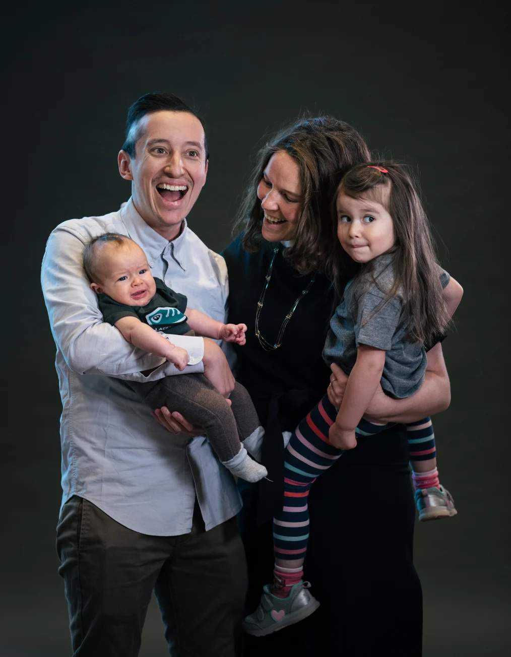 This picture shows a family of four with medium-skin tones smiling and looking
towards the camera, with a dark gray background.