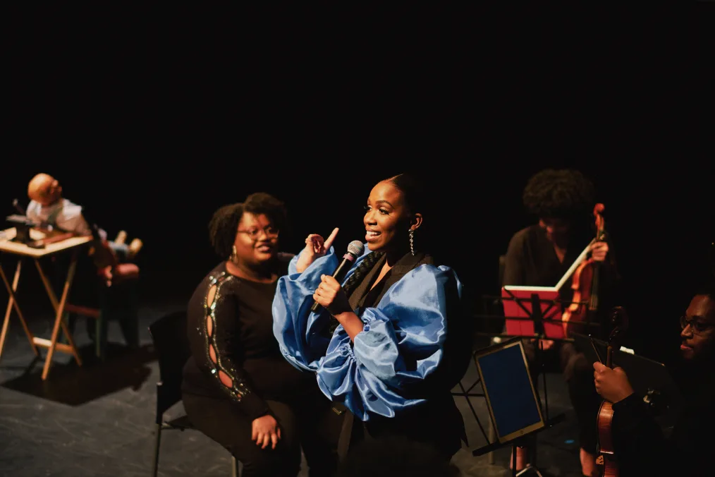 Kori Coleman, a Black woman with a deep skin tone stands on a stage and holds a microphone. She stands in front of a string quartet of Black people with a range of medium to deep skin tones. Her index finger is raised.