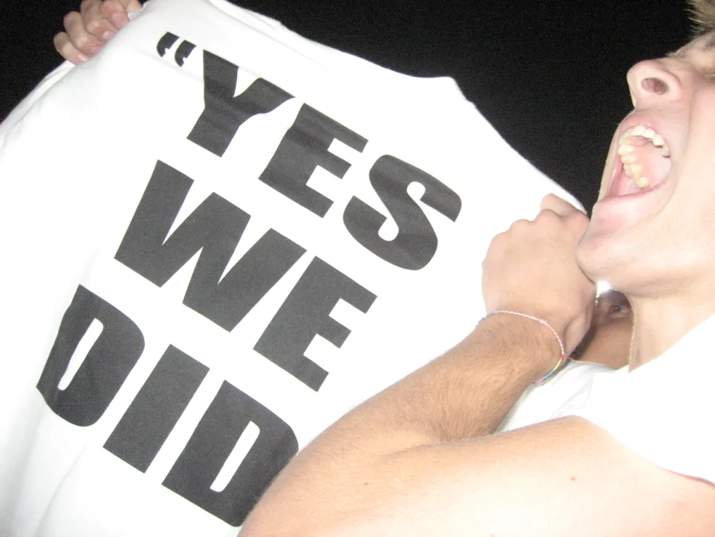 A person with their mouth open is cropped partially out of the photo. They hold up a white t-shirt with the words "YES WE DID" in bold, black letters.