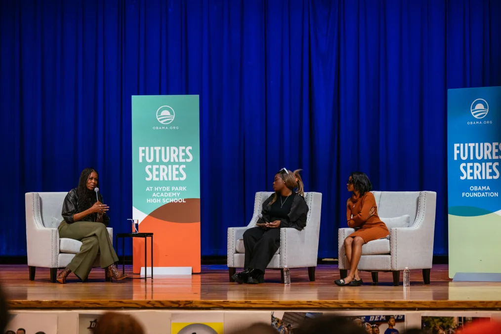 Young people fill a large auditorium. Mrs. Obama sits in a light gray chair next to two young women, also seated.