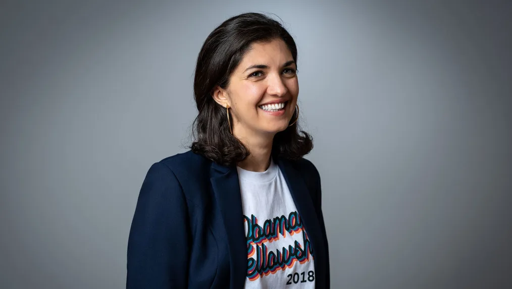 A woman with shoulder-length dark hair looks off to the right. She wears hoop earrings and a suit jacket over a t-shirt with the words "Obama Fellows 2018" on it.