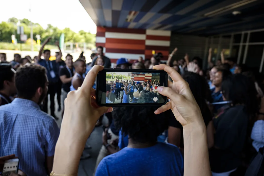 Two hands with a light skin tone hold an iPhone sideways recording a group of individuals with a variety of skin tones dancing.  