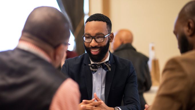 A Black man with short black hair and full beard wears glasses, bow tie and suit jacket. He is smiling. In front of him we see the backs of two heads.