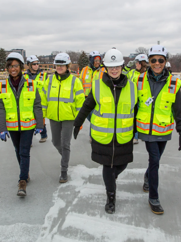 Valerie Jarrett smiles and walks leading a crowd of people with various skin tones at a construction site. All are wearing pants, coats, gloves, and jackets with yellow safety vests and white hard hats. 