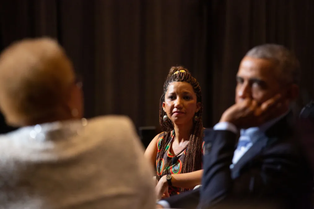 

The camera focuses on a woman with a deep medium skin tone with brown braids as she looks at President Obama and a woman with a medium skin tone  