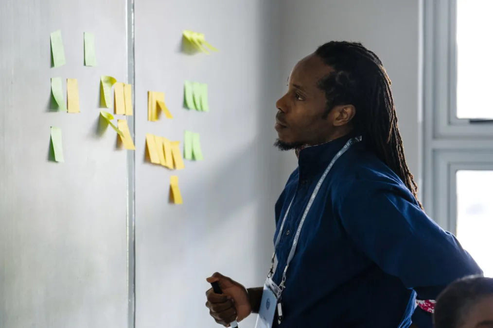 A black male with a deep skin tone and long locs looks at sticky notes on the wall in the colors of yellow and a lighter green.