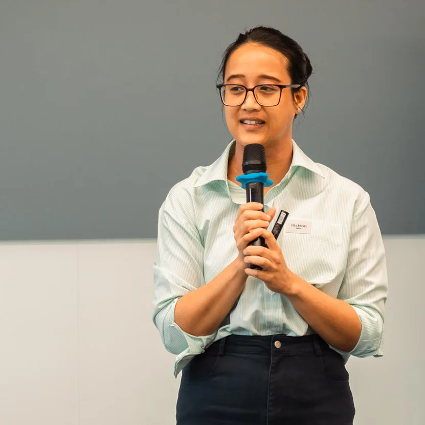 Viria, a woman with a medium skin tone and short black hair, holds a microphone as she speaks to an audience. She is wearing glasses. In the background, a poster reads, “intention.”