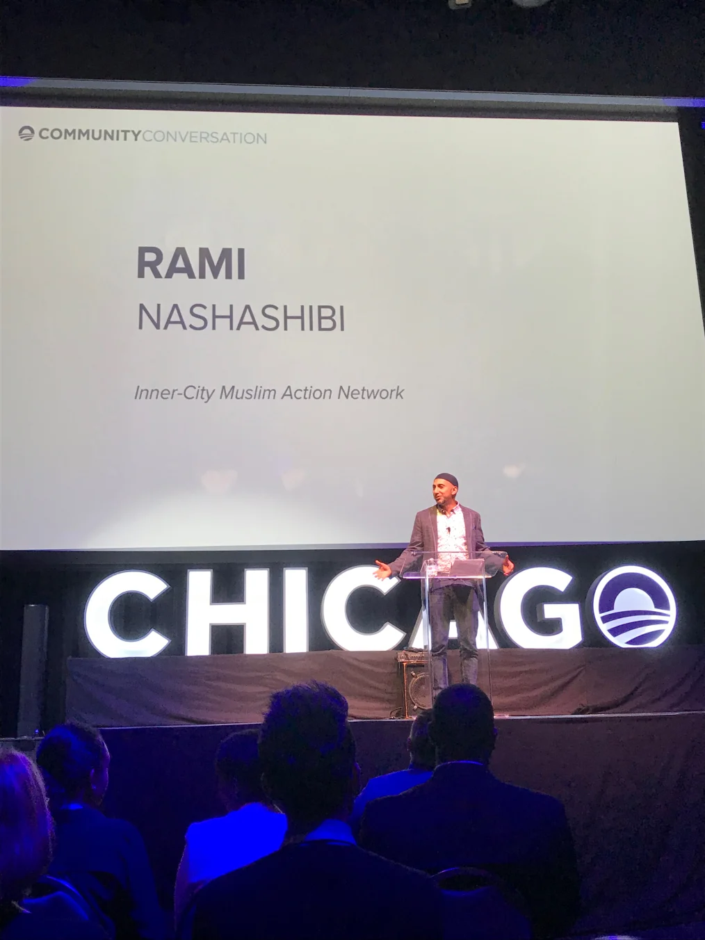 A man with a light medium skin tone dressed in business attire stands on stage in front of a clear podium. Behind him is a large white screen with text that reads "Community Conversation" and below it, the text reads "Rami Nashashibi" and "Inner-City Mulsim Action Network" 
