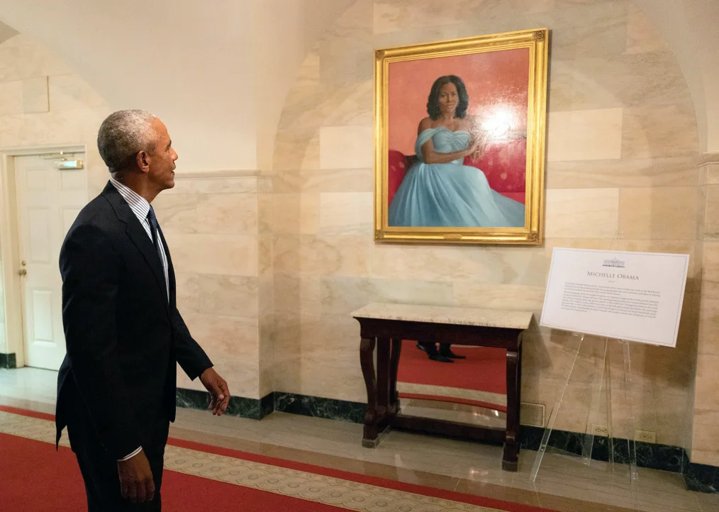 President Barack Obama wearing a black suit stands on the left-hand side of the photo and looks at a painting of First Lady Michelle Obama on a beige wall. 
