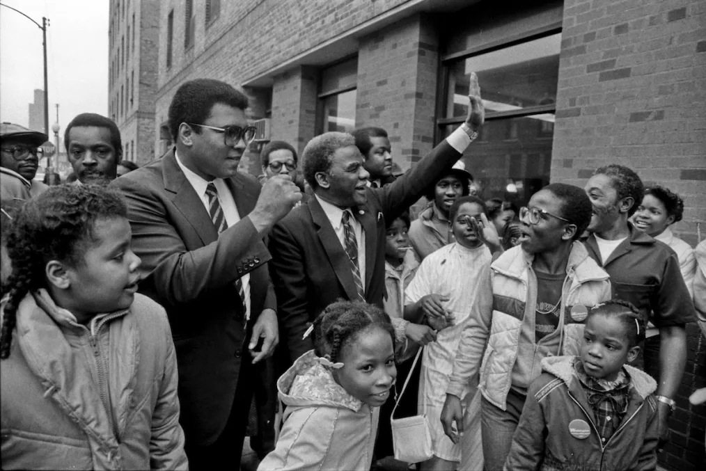 A black-and-white image shows Muhammed Ali wearing glasses and with his fist in the air, surrounded
by young children with deep skin tones.