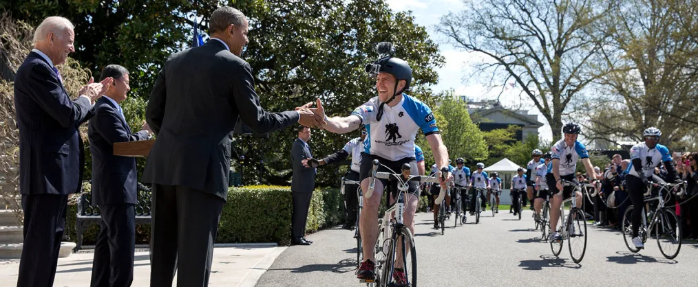 President Obama shakes the hand of a male biker with a light skin tone wearing black shorts, a blue and white shirt, and a black helmet. There are a group of bikers behind the biker and there are two individuals with black suits standing 