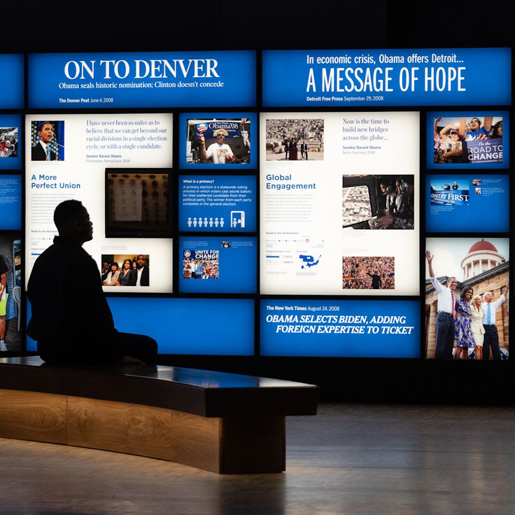 A man in shadows sits on a bench in front of two digital screens at the Obama Presidential Center Museum. 
