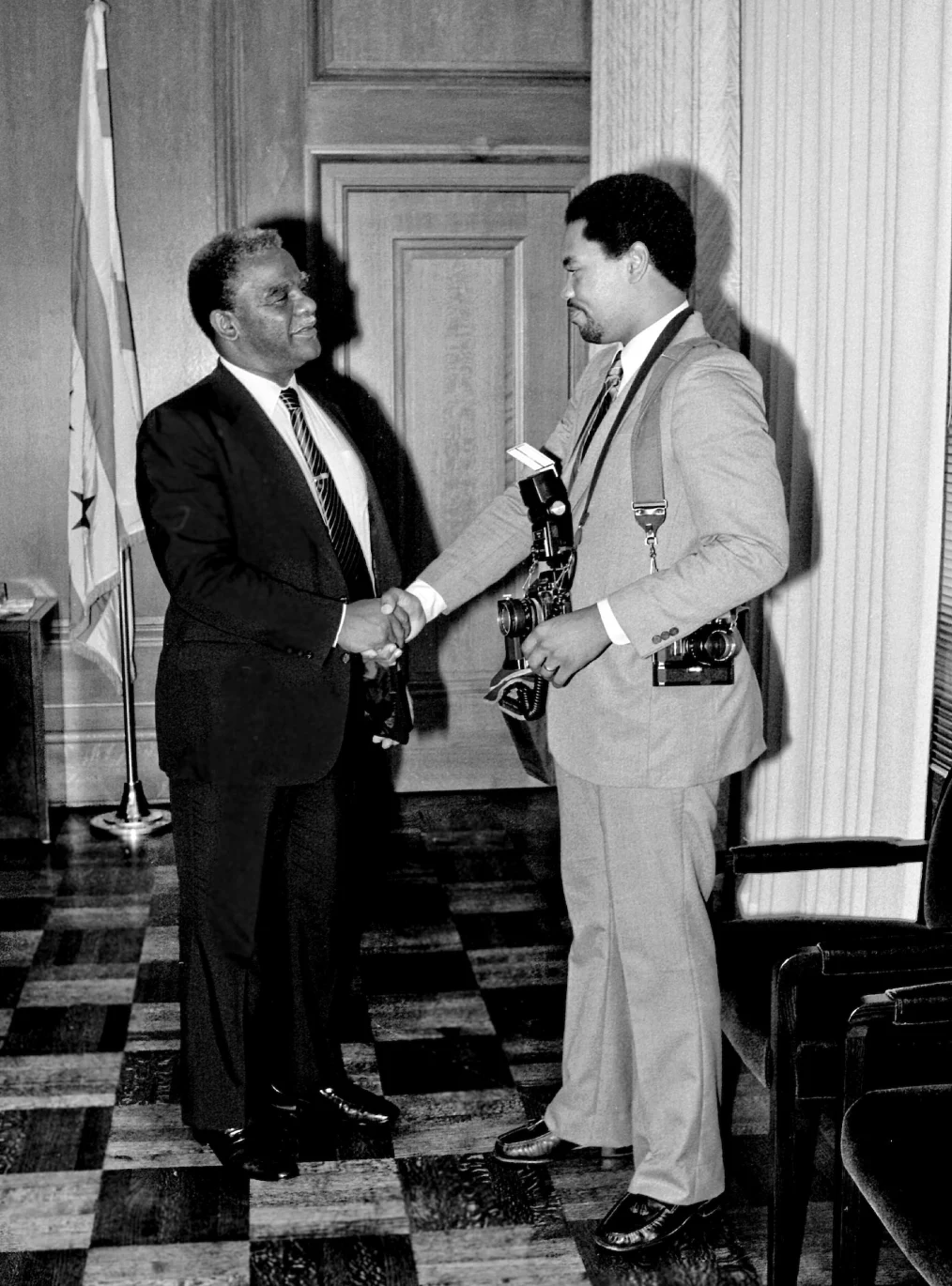 A black and white photograph of a young Black photographer carrying two film cameras shakes hands with Mayor Harold Washington.