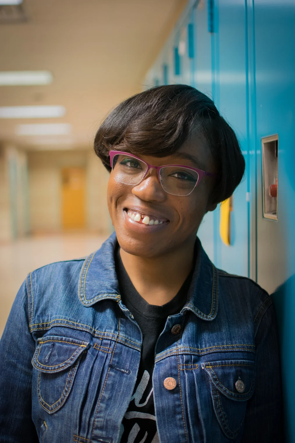 A deep skin toned Black woman with ear length dark hair is smiling with her teeth showing. She is wearing pink framed glasses, a black T-shirt and a denim jacket. She is leaning to the right against a set of blue lockers in a school hallway.