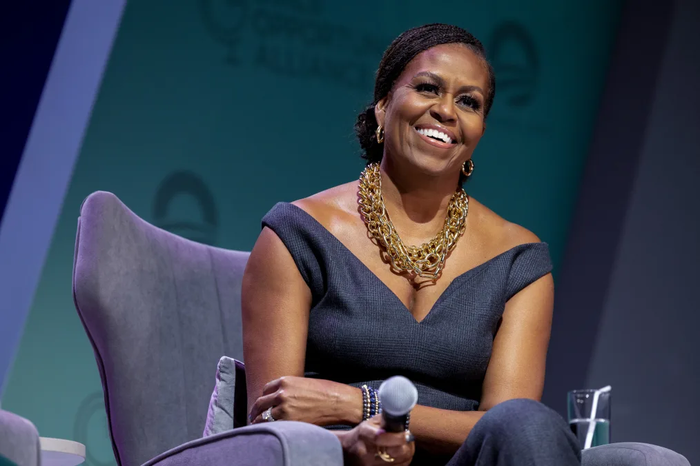 Mrs. Obama smiles on stage holding a microphone. She wears a chain necklace and a dark gray top. 