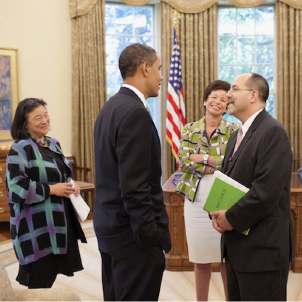 A image of Brian Bond standing in the Oval Office with President Obama, Tina Tchen, and Valerie Jarrett. Brian has an olive complexion and closely cropped dark hair, and glasses. He is wearing a dark colored suit, while facing President Obama. In his hands is a green memo. President Obama has a medium deep complexion and black and gray hair. He is wearing a dark colored suit and facing Brian Bond with his hands in his pocket. Tina Tchen has a medium olive complexion and dark hair cut to her shoulders. She is smiling at President Obama and Brian Bond. She is wearing a black dress with a plaid overcoat. Valerie Jarrett has a light deep complexion, short brown hair, and glasses. She is smiling in the direction of President Obama and Brian Bond with her arms crossed against her chest. She is wearing a green jacket and a light color skirt.