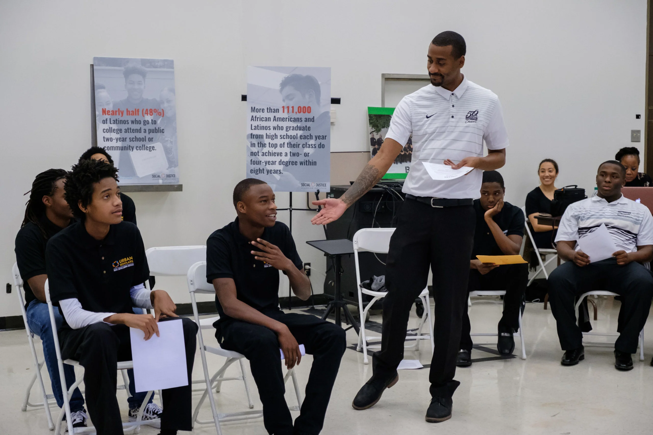 A seated group of young people apart of Urban Scholars, with medium to deep skin tones, have a meeting. A middle age man with medium-deep skin tones leads the group. He is standing, has short hair and has a sideburn shaved box beard. He wears a white nike polo with blue stripes, a black belt, with black pants, and boots. His right arm is extended towards a young man. In the background are two posters that share statistics regarding "minorities" and their college achievements.