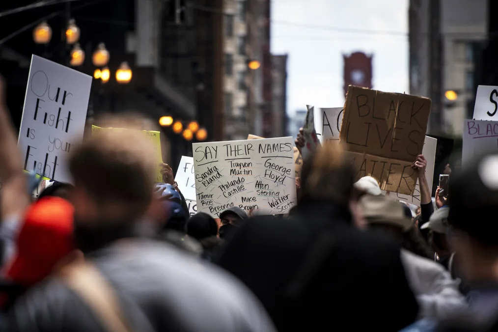 An image of a large group of various people outdoors holding cardboard and paper signs that reads "Black Lives Matter" "Our fight Is Your Fight" and "Say Their Names"
