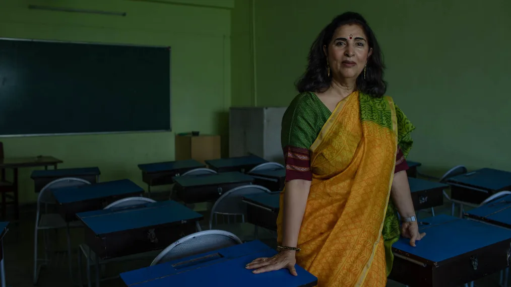 A older women with long brown hair, medium skin with warm undertones, a red dot with three small dots in a pryamid shape above just below her forehead, and rosy cheeks. She is wearing fabrics typically seen from south-asian cultures. She is standing with her hands on two school desks. She is in a green classroom with a chalk board in the backgrond. The room is dark and in ok condition. 