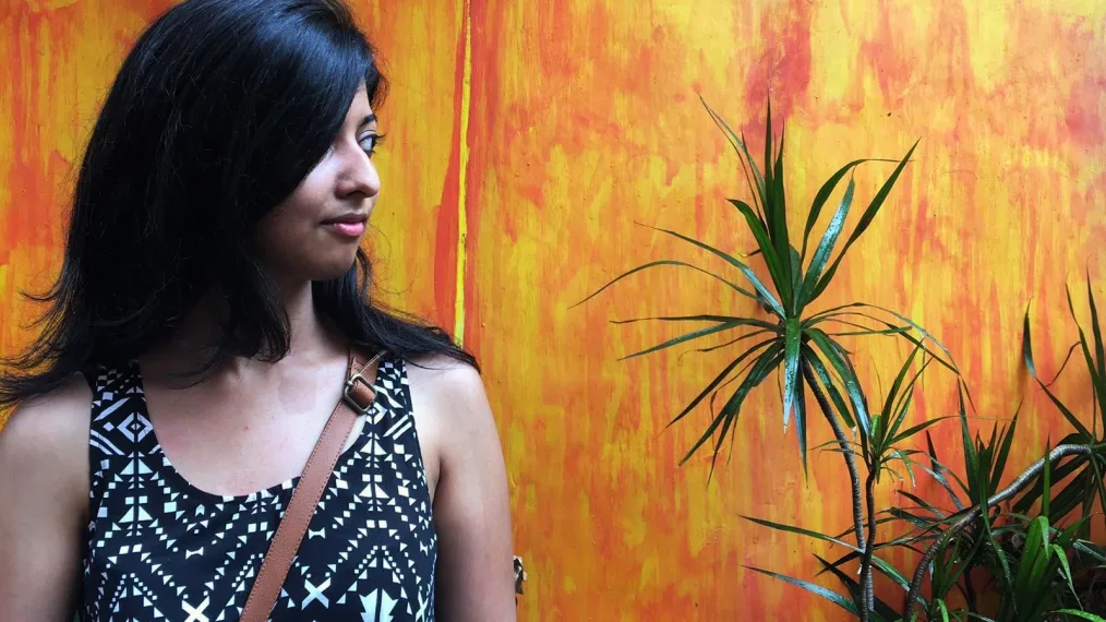 A woman with medium-dark skin tone and shoulder-length, straight black hair, looks to the right. She stands against a bright orange backdrop. A green plant is beside her.