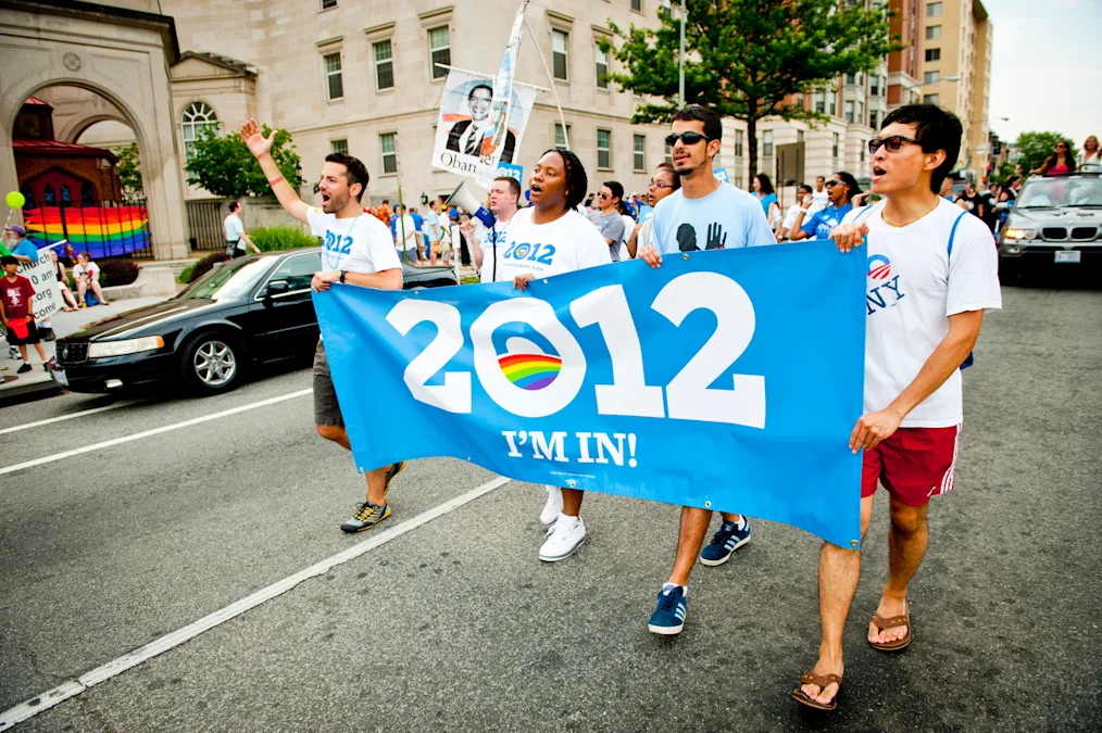 Four people with a range of light to deep skin tones walk behind a banner at a parade that reads, “2012” and “I’m in.” All are wearing shirts with the Obama logo inside the 2012. More people walk behind them in the background.