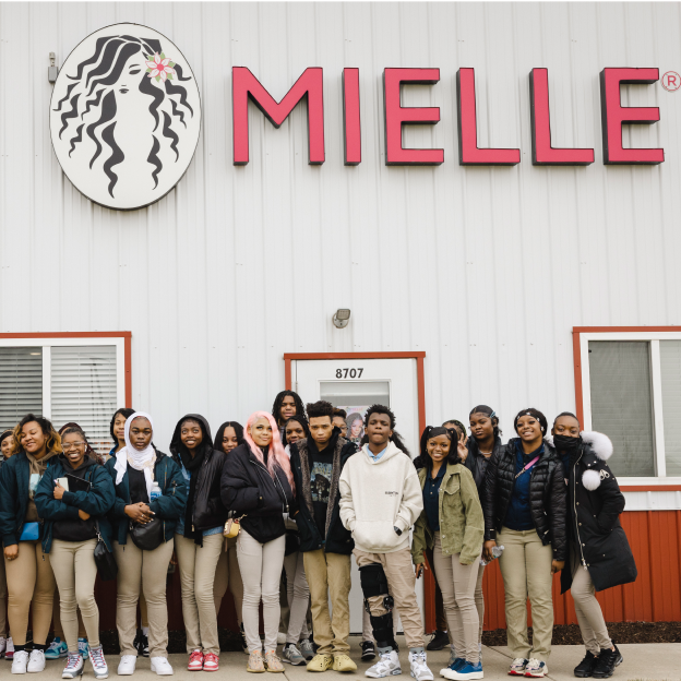 A group of students with medium-light to medium-dark skin tones, wearing khaki pants and a variety of hair styles, stand in front of a white building that reads “Mielle” and a graphic of a woman with long black hair on the wall.