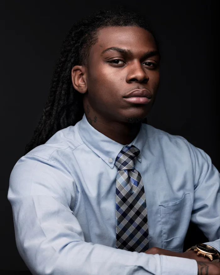 A portrait photo of a young man with a deep skin tone wearing a light blue button-up shirt and black, gray, and blue tie in front of a gradient black background. 