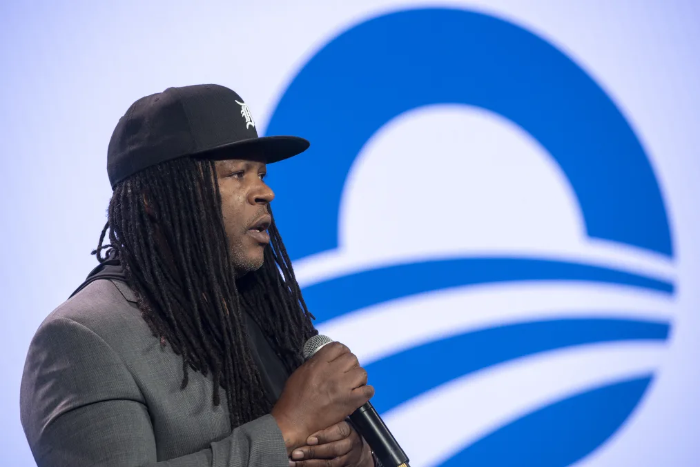 In this portrait, a black man with a deep skin tone and locs wearing a grey suit jacket and black baseball cap holds a microphone.