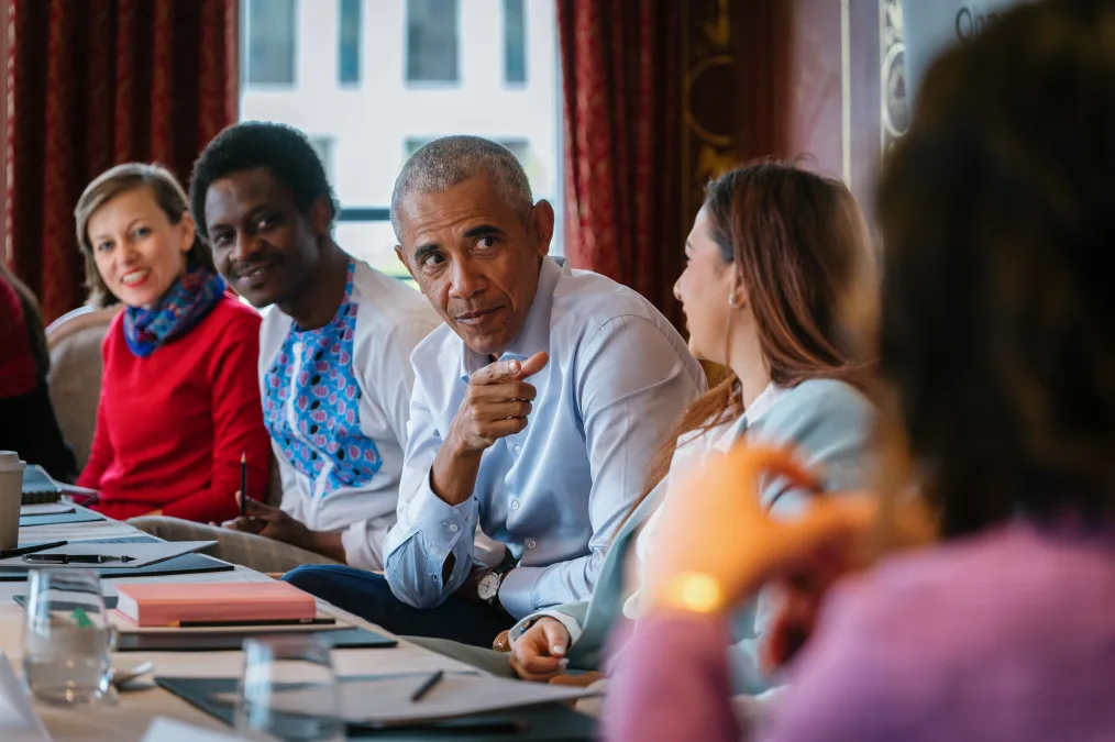 President Obama sits next to three people with a range of light to deep skin tones at a wooden table. He is pointing at a young woman wearing a light blue blazer who looks at him smiling.
