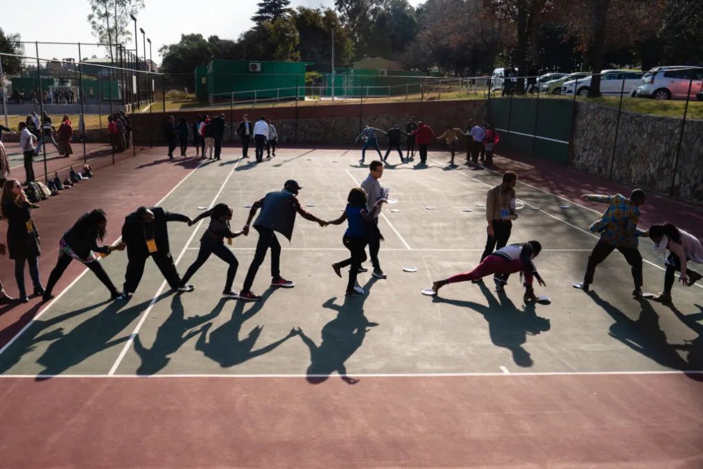 A group of people with various skin tones, holding hands, and step on white paper plates on a concrete tennis court.