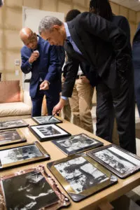 The camera focuses on President Obama in a suit looking over black and white photos laying on a table. Surrounding him are men with skin tones ranging from medium to dark, also in business attire. 