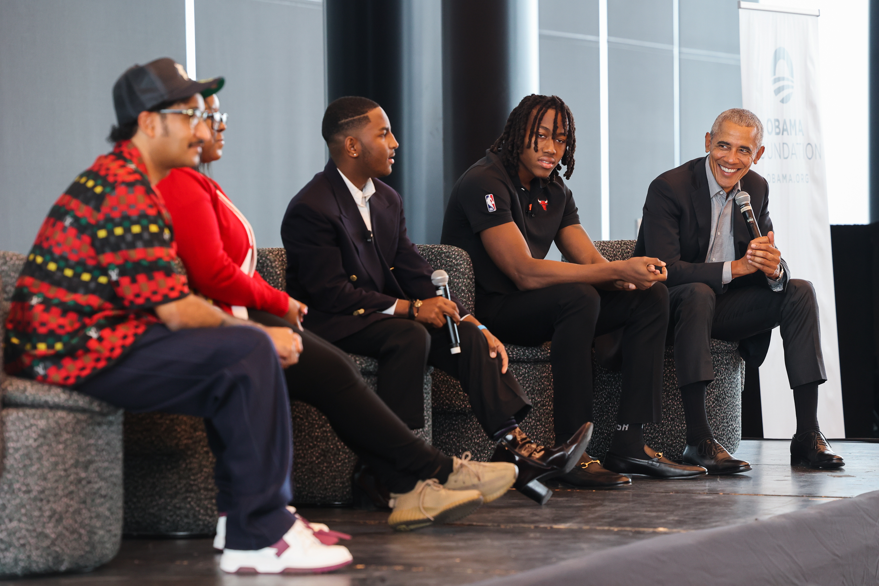 Four young people in business and casual attire sit on stage in chairs while President Obama, also seated, smiles holding a microphone.