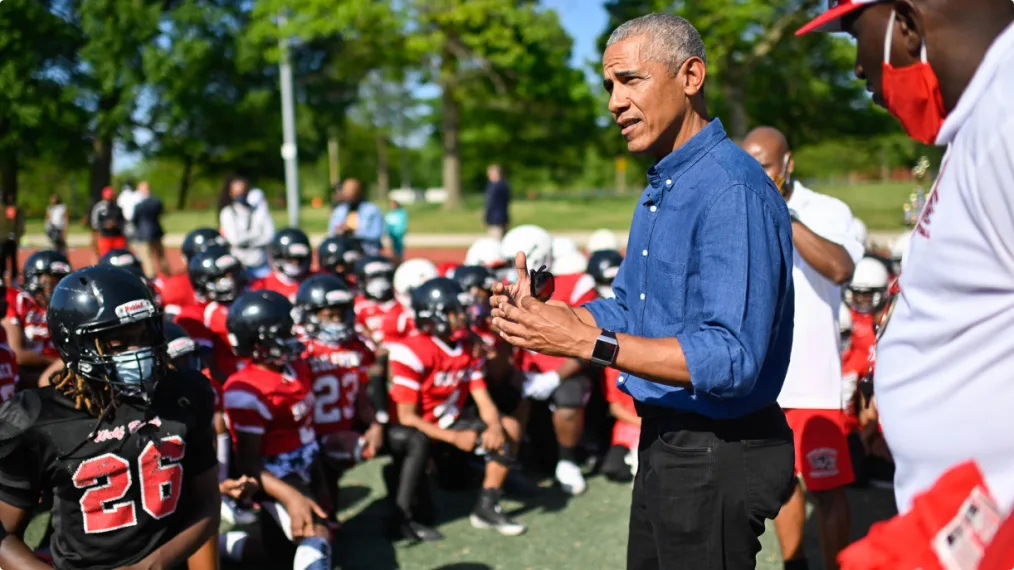 Barack Obama squints slightly  and gestures with his hands as he looks out over a team of football players wearing red and black jerseys. It is very sunny and there are green trees behind them. 
