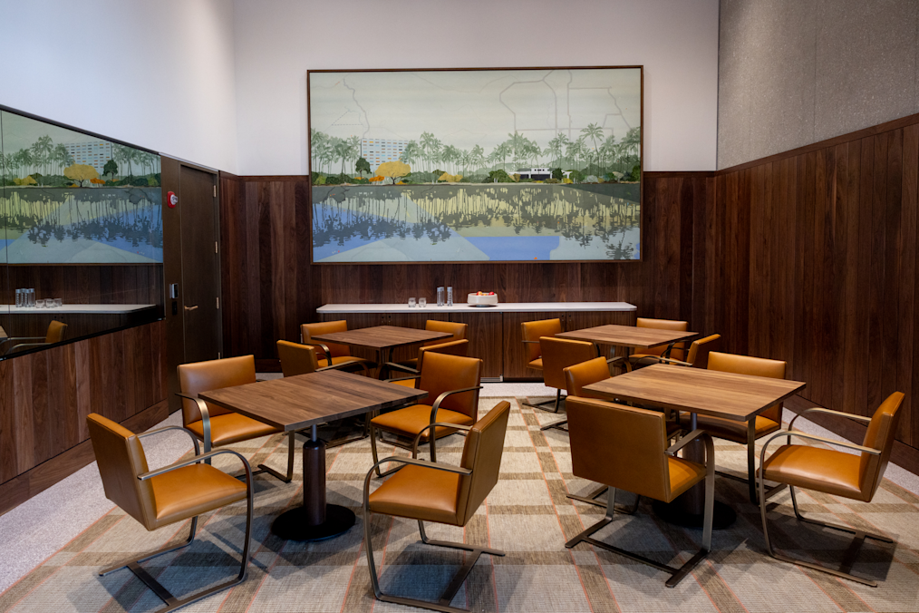 In a room with dark wood walls, four square wooden tables each have four tan leather armchairs around them. A large painting of palm trees hangs on the back wall above a white counter. On the left wall, a large mirror reflects the room.