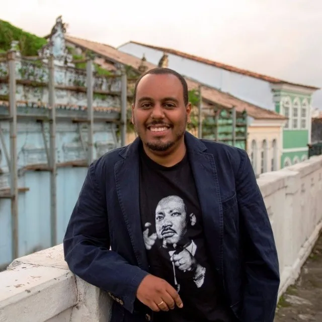 A man with medium-dark skin tone smiles at the camera. He has close-cropped hair, mustache and a short beard. He wears a t-shirt with a photo of Martin Luther King, Jr. on it and a blue suit jacket. Behind him appear red-tile rooftops of buildings.