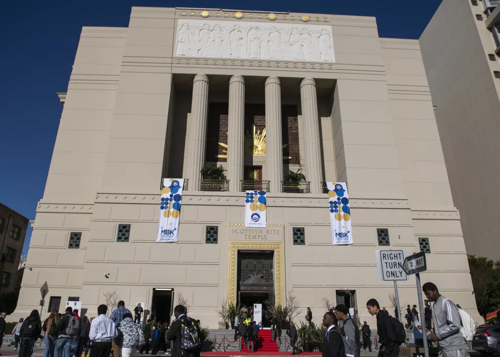 The Scottish Rite Temple, a large beige stone buliding with columns and gold accents, draped with ¨MBK Rising¨ has various visitors surrounding the outside.
