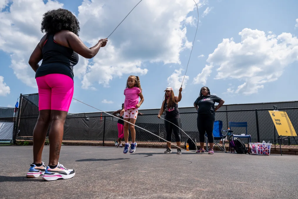 A low angle shot of five girls and women jumping double dutch. A young girl with a light skin tone jumps in the middle. Two Black women with deep skin tones hold the rope. A Black woman with a deep skin tone watches on the sidelines. Another woman with a deep skin tone watches from a distance. A yellow sign in the background reads, “jump rope.” 