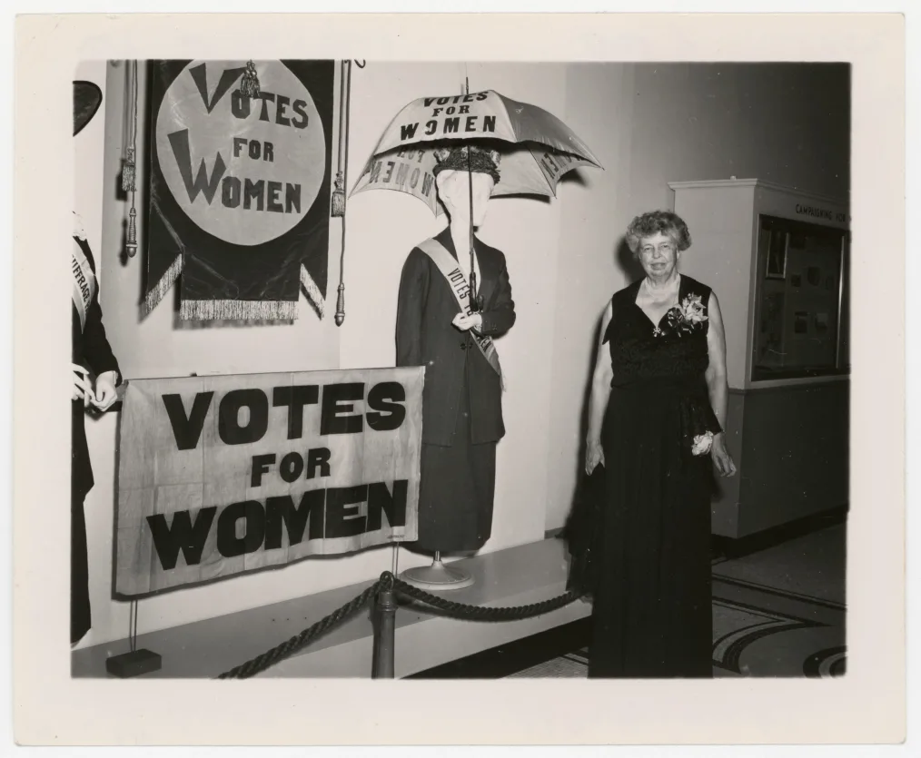 
A black and white photo of an older woman with a light skin tone stands grinning in front of multiple posters that read: “Votes For Women”