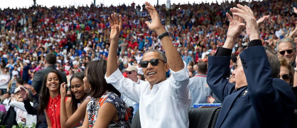 President Obama is wearing sunglasses and a white collared shirt raising his arm toward the camera. The background includes the first family and a large crowd behind them.