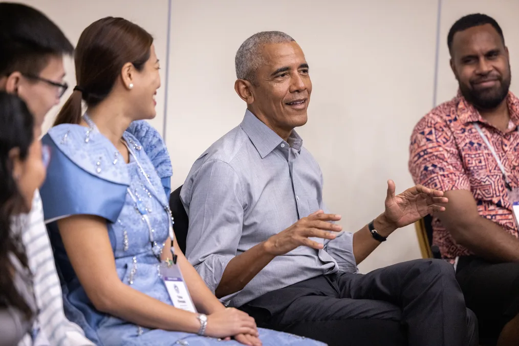 President Obama sits in a circle and speaks to Leaders with a range of ages and light to medium skin tones. His arms are outstretched. He is wearing a gray button up. 
