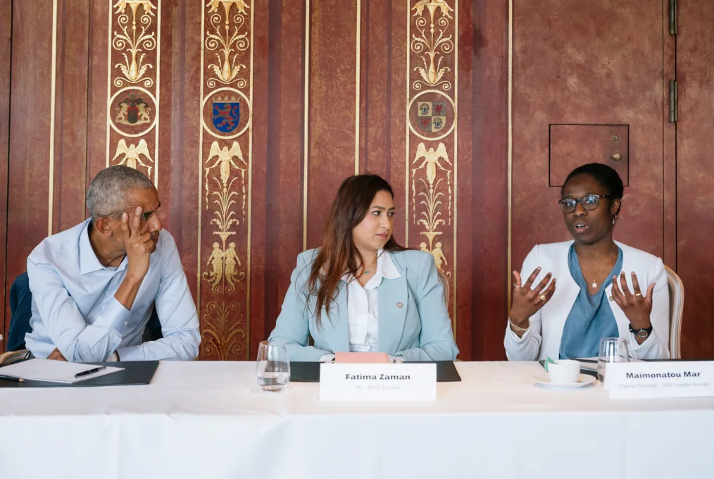 President Obama rests two fingers on his forehead as he listens to Maïmonatou Mar, a Black woman with a deep skin tone, speak. In the middle of the two is Fatima Zaman, a woman with long brown hair and a light medium skin tone.