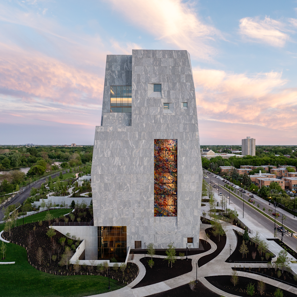 A high-angle photo shows the Obama Presidential Center Museum which is a tall light gray stone building on a green lawn during sunset. The building has an uneven shape with small windows. A tall rectangle filled with colorful stained glass sits near the center of the tower. At the bottom left, large glass windows light up an entrance. Smooth concrete paths curve around black dirt gardens and young green trees in front of the building. On the right, a multi-lane street with cars runs alongside a row of buildings and trees. In the background, green trees stretch toward the horizon under a soft pink and blue sky with light clouds.

