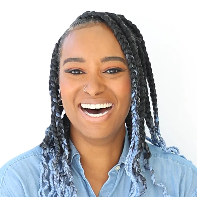 A headshot of a woman with medium-dark skin tone smiling widely, her mouth open and teeth showing. Her black hair is in braids, fading to blue at the ends. She wears a light blue collared shirt.